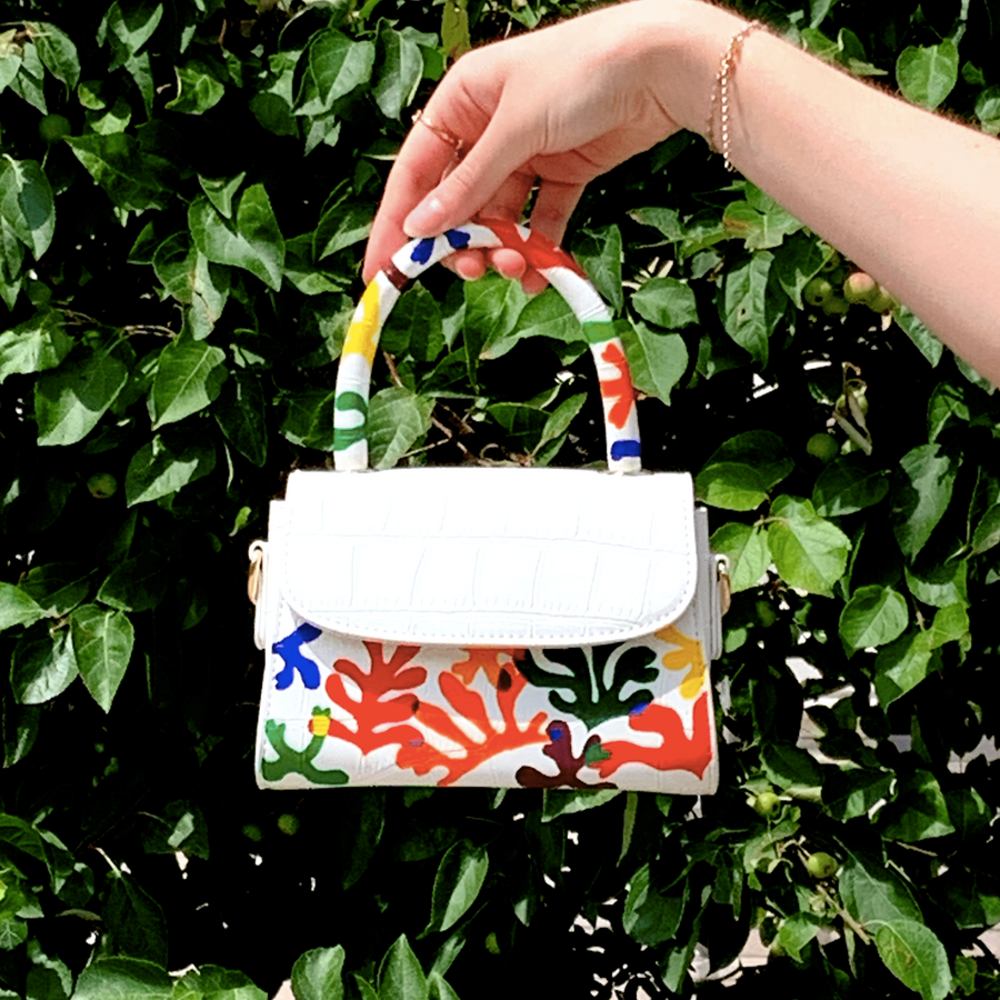 Hand holding a white hand-painted Matisse inspired bag in front of a wall of leaves.